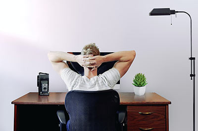 Ein Mann sitz mit hinterm Kopf verschränkten Armen an einem Schreibtisch.