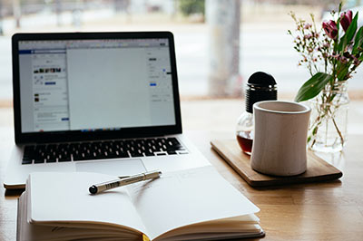 Ein Laptop, Notizbuch, Kaffeetasse und eine kleine Vase mit Blumen stehen auf einem Tisch. 