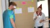 Gruppe Mitarbeitenden steht vor einer Papierwand mit der Aufschrift "Krisenmanagement" und führt etwas mit Gebastelten Gegenständen vor.